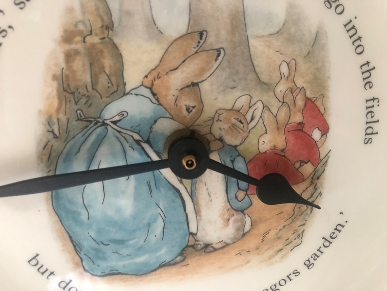 A close up of the black clock hands and the image of Mrs Rabbit fastening Peter Rabbits blue jacket. The three Flopsy bunnies in their red capes are in the background on their way towards the woods. Mrs Rabbit wears a blue dress and white apron