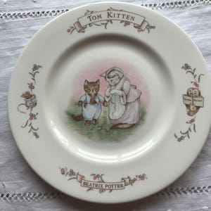 Puede incluir: Plato de cerámica blanco con una ilustración de dos gatos de la historia de Tom Kitten. El plato tiene un centro rosa y está decorado con motivos florales y los textos "Tom Kitten" y "Beatrix Potter".