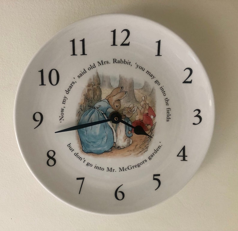 A white ceramic circular clock in the style of a plate. Black numerals and hands with central image an illustration by Beatrix Potter of Mrs Rabbit doing up Peter Rabbits blue jacket and the three Flopsy bunnies in the background in their red capes.