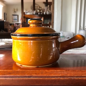May include: A brown ceramic pot with a lid and a single handle. The pot has a black rim and a glossy finish.