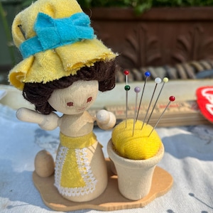 May include: A wooden figurine with brown yarn hair wearing a yellow fabric hat with a blue bow and a yellow apron with white lace trim. The figurine stands on a base next to a small wooden pot containing a yellow pincushion with colourful pins.