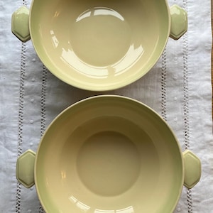 May include: Two pale yellow ceramic bowls with green rims and octagonal handles. The bowls are empty and sitting on a white tablecloth.