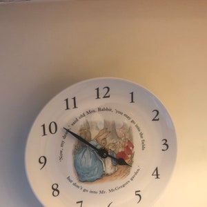 A white ceramic circular clock in the style of a plate. Black numerals and hands with central image an illustration by Beatrix Potter of Mrs Rabbit doing up Peter Rabbits blue jacket and the three Flopsy bunnies in the background in their red capes.