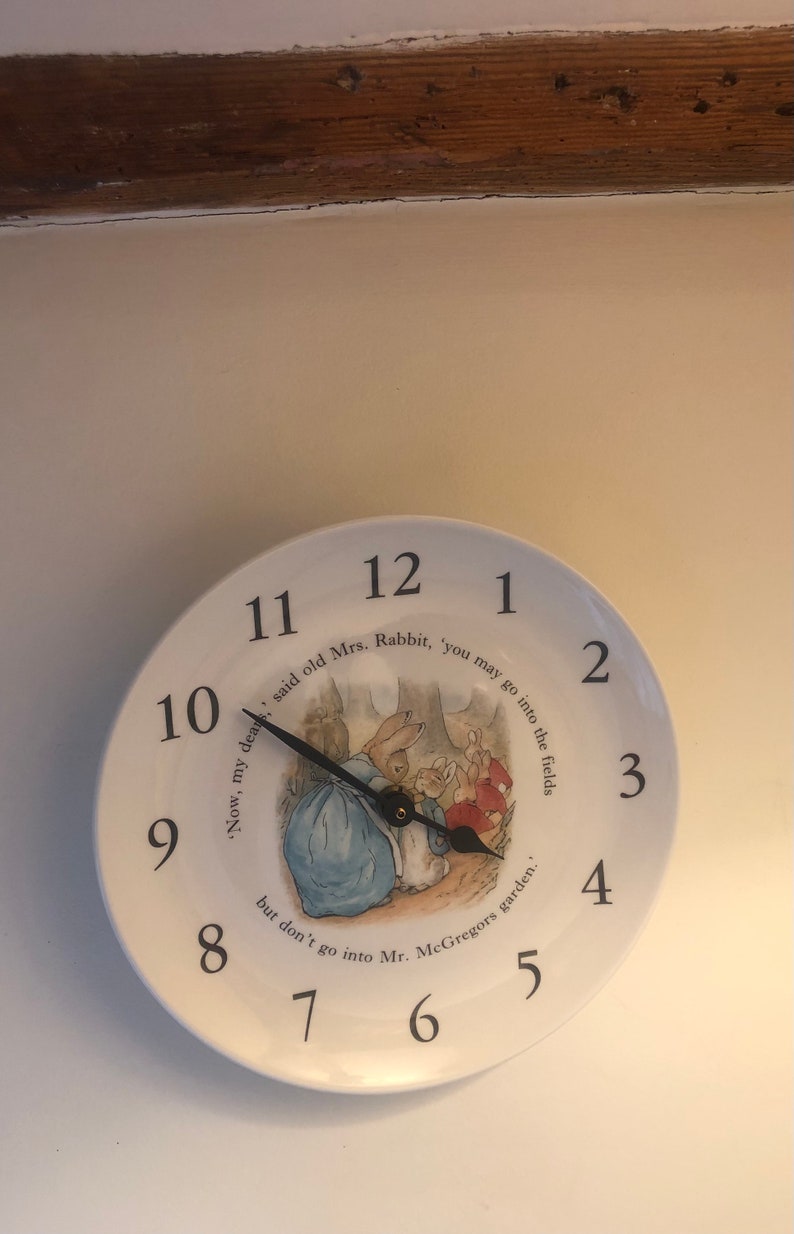 A white ceramic circular clock in the style of a plate. Black numerals and hands with central image an illustration by Beatrix Potter of Mrs Rabbit doing up Peter Rabbits blue jacket and the three Flopsy bunnies in the background in their red capes.