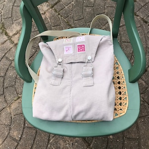May include: A light beige corduroy backpack with adjustable straps and small embroidered patches. The backpack is sitting on a green chair with a woven seat.