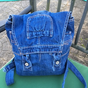 Könnte beinhalten: Ein blauer Jeans-Rucksack mit einer Klappe und zwei Knopfverschlüssen. Der Rucksack hat einen langen verstellbaren Riemen.