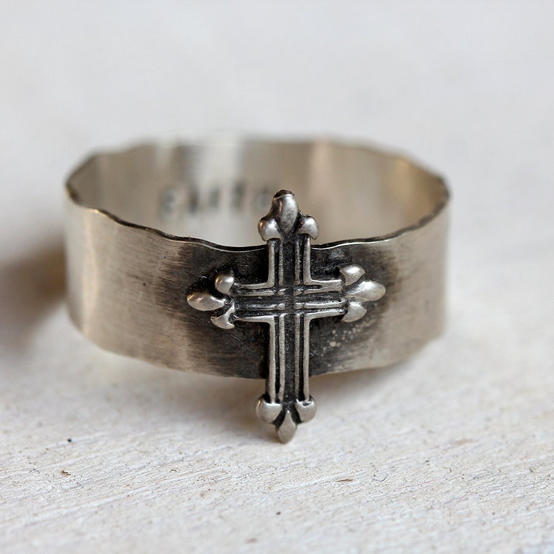 May include: A silver ring with a cross design. The cross has a fleur-de-lis design at the top and bottom.