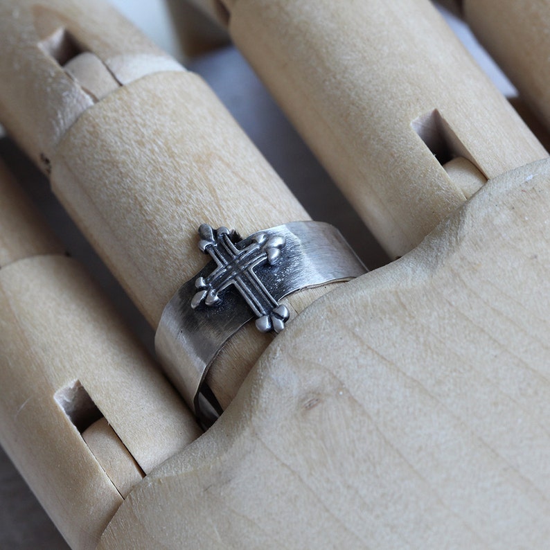 May include: A silver ring with a cross design. The ring is on a wooden hand model.