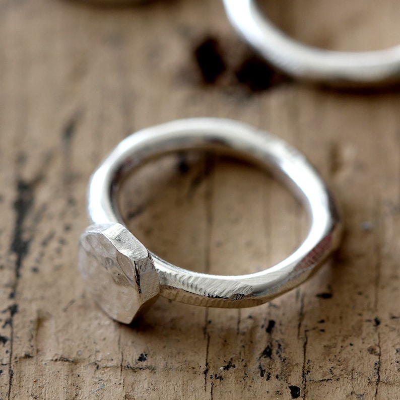 May include: A silver ring with a hammered, octagonal band. The ring is made of metal and has a simple, modern design.