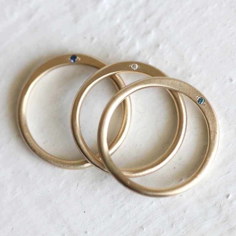 May include: Three gold rings with a small blue gemstone on the left ring, a small white gemstone on the middle ring, and a small green gemstone on the right ring.