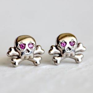 Puede incluir: Un par de pendientes de plata con forma de calavera y huesos cruzados con detalles de gemas rosas.