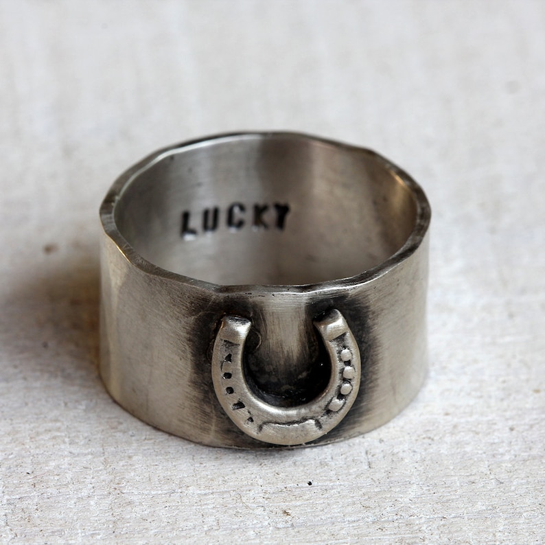 Lucky horseshoe ring from Praxis Jewelry. Sterling silver wide band ring with a horseshoe on the front and the word lucky engraved on the inside of the hammered band.