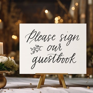 May include: A white sign with black calligraphy that reads "Please sign our guestbook" with a small floral design. The sign is on a wooden easel and is sitting on a white table with gold confetti.