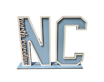 May include: Wooden sign with the letters NC in blue with a white outline. The word "north carolina" is written vertically on the left side of the N.