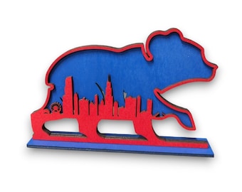 May include: A wooden Chicago bear figurine with a red silhouette of the Chicago skyline inside. The bear is blue and red and standing on a wooden base.