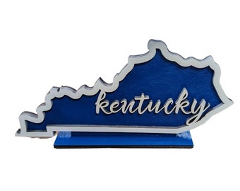 May include: A blue wooden cutout of the state of Kentucky with a white outline and the word "Kentucky" written in white lettering.