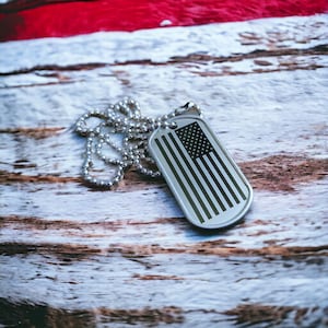 May include: A silver dog tag with a black and white American flag design on a silver chain. The dog tag is lying on a weathered wooden surface.