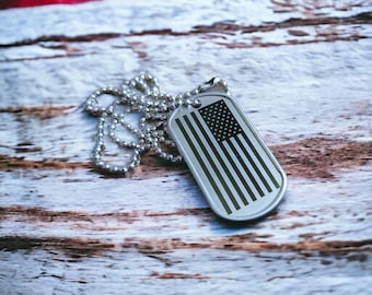 May include: A silver dog tag with a black and white American flag design on a silver chain. The dog tag is lying on a weathered wooden surface.