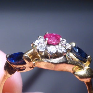May include: A gold ring featuring a central cluster of small white diamonds surrounding a round, red ruby. Two dark blue, marquise-cut gemstones flank the central cluster. The ring's band is a simple, polished gold.