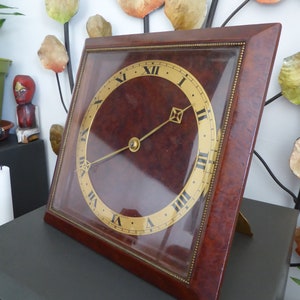 May include: A square mantel clock with a dark brown burl wood frame and a glass front. The clock face is circular, featuring a gold-toned background with black Roman numerals and thin, gold-colored metal hands. The clock is angled on a stand.