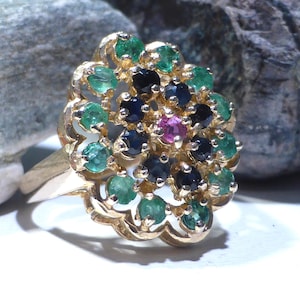May include: A gold ring featuring a floral-shaped cluster setting. The centre is a single pink gemstone surrounded by a ring of dark blue sapphires, with an outer border of green emeralds set in a scalloped, openwork gold metal frame.