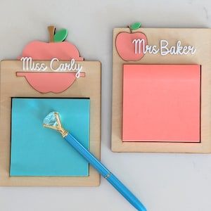 Puede incluir: Dos porta notas de madera con diseños de manzanas y nombres personalizados. El porta notas de la izquierda dice "Miss Carly" y tiene una nota adhesiva azul y un bolígrafo azul con una parte superior de pedrería. El porta notas de la derecha dice "Mrs. Baker" y tiene una nota adhesiva rosa.