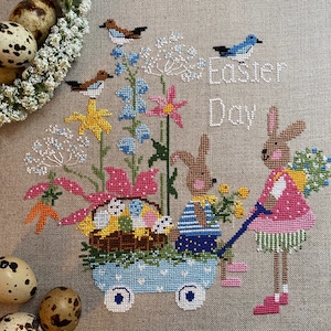 Può includere: Ricamo a punto croce con due conigli, uno che tira un carrello pieno di uova di Pasqua e fiori, l'altro che cammina al suo fianco. Il disegno include il testo "Easter Day" e diversi uccelli.