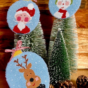 Könnte beinhalten: Drei blaue und weiße Weihnachtskugeln mit verschiedenen Designs. Die erste Kugel zeigt einen Weihnachtsmann mit roter Mütze und Bart. Die zweite Kugel zeigt einen Schneemann mit grünem Hut und rotem Schal. Die dritte Kugel zeigt ein Rentier mit braunen Geweih und einer schwarzen Fliege.