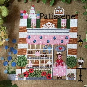 May include: A cross-stitch embroidery depicting a French patisserie with a pink awning, a window display of pastries, and a woman in a pink dress standing outside. The shop is decorated with flowers and a sign that reads "Patisserie".