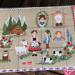 Può includere: Un ricamo a punto croce che rappresenta una scena alpina tradizionale con una casa, montagne, fiori e figure in abiti tradizionali. Il ricamo è incorniciato da un bordo in pizzo bianco e un nastro a quadri rossi e bianchi.