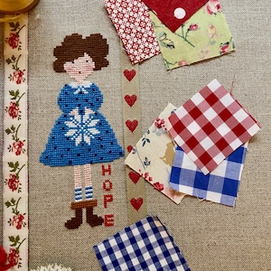 Può includere: Ricamo a punto croce di una ragazza con un vestito blu con un motivo a fiocco di neve. La parola "HOPE" è ricamata sotto di lei. L'immagine include diversi pezzi di stoffa in motivi a quadri rossi, bianchi e blu, stampe floreali e pois.