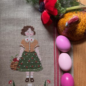 Può includere: Un ricamo a punto croce di una ragazza che tiene un cesto di fiori, con un fiore rosso e tre uova rosa su uno sfondo di lino.