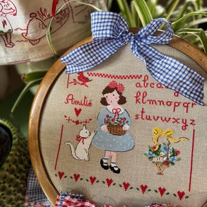 Könnte beinhalten: Ein Kreuzstich-Stickrahmen mit einem Mädchen, einer Katze und dem Alphabet-Design in roter, blauer und schwarzer Farbe auf beigem Stoff. Oben ist eine blau-weiß karierte Schleife angebracht. Der Rahmen ist aus Holz.
