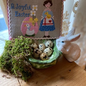 Può includere: Un ricamo a punto croce con il testo "A Joyful Easter" e una ragazza che tiene un pulcino giallo. Il ricamo è incorniciato da un bordo rosa ed è esposto su un tavolo di legno con un nido di muschio e uova di quaglia.