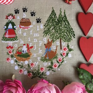 Può includere: Ricamo a punto croce con una ragazza in un vestito rosso, una volpe e un cestino di latte. L'alfabeto è ricamato attorno al bordo del disegno, con una scena di bosco sullo sfondo. Il ricamo è circondato da rose rosa e cuori rossi.