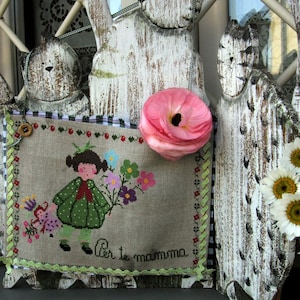 Può includere: Un telaio in legno bianco per ricamo a punto croce con un disegno a punto croce bianco, verde e rosso di una ragazza che tiene dei fiori. Il testo "Per te mamma" è ricamato nella parte inferiore del disegno. Un fiore rosa è sul lato destro del telaio.