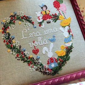 Può includere: Un ricamo a punto croce incorniciato con una ghirlanda a forma di cuore di foglie verdi, bacche rosse e fiori bianchi. Il testo "Con un alma  santa" è ricamato al centro del cuore. Il ricamo presenta una ragazza con un libro, un uccello e due conigli.