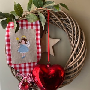 May include: A wicker wreath decorated with a red and white gingham banner featuring a cross-stitched angel, a red heart ornament, and a red and white ornament.