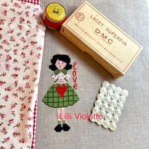 Può includere: Ricamo a punto croce di una ragazza che tiene un cuore con la parola "Love" sopra di lei. Il ricamo è su un tessuto di lino beige con il testo "Lilli Violette" sotto la ragazza. L'immagine include anche un rocchetto di filo rosso, una scatola di filo da ricamo DMC e un pacchetto di bottoni bianchi.
