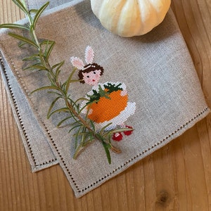 Può includere: Un tovagliolo di lino grigio con un ricamo a punto croce di una ragazza che tiene una zucca. Il tovagliolo è piegato e ha un bordo decorativo. Una zucca bianca è appoggiata sul tovagliolo.