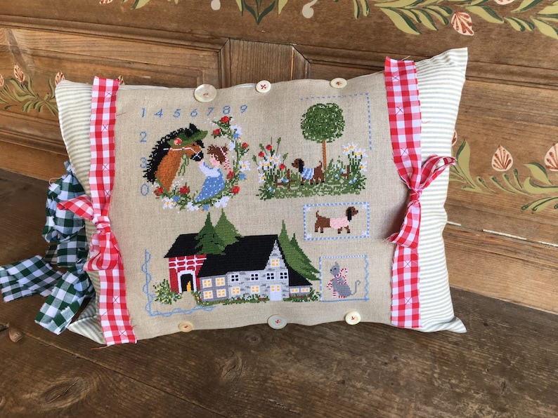 May include: A beige pillow with red and white gingham ribbon trim. The pillow features a cross-stitch design of a girl with a horse, a house, a tree, and several dogs. The numbers 1 through 9 are stitched on the pillow.