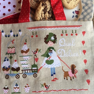 Puede incluir: Una bolsa de lona beige con un diseño de punto de cruz que presenta a una niña con un perro, cupcakes y el texto "Quel Delice!" La bolsa tiene asas rojas y un forro de tela de lunares rojos y blancos.