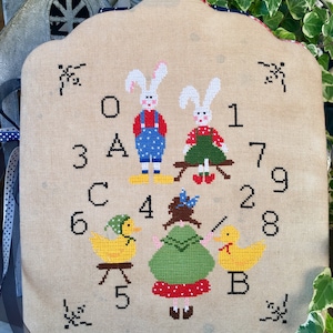 May include: A cross-stitch wall hanging with a playful design of bunnies, ducks, and a girl, plus numbers and letters. The figures are in bright colours, including red, green, and yellow, on a beige background. The edges are scalloped.
