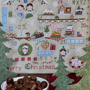Può includere: Un calendario dell'Avvento natalizio ricamato, con immagini di bambini, prodotti da forno e decorazioni festive. Il testo "Merry Christmas" è ricamato in basso. Una ciotola di castagne e alberi di Natale in feltro in primo piano.