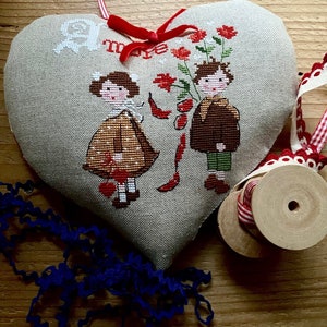 Può includere: Un cuscino a forma di cuore con un ricamo a punto croce che raffigura due bambini, una bambina e un bambino, che tengono dei fiori. Il cuscino è in tessuto beige e ha un nastro rosso legato intorno. Lo sfondo è una superficie di legno.