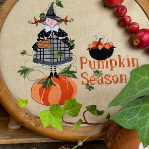 Può includere: Un ricamo a punto croce incorniciato con una strega che tiene un cestino, in piedi su una zucca. Il testo "Pumpkin Season" è ricamato sotto la strega. La cornice è circondata da edera verde e mele rosse.