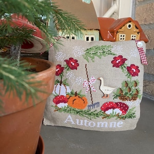 May include: A beige pillow with a cross-stitch design featuring a goose, pumpkins, mushrooms, and the word "Automne". The pillow is sitting on a windowsill with small ceramic houses in the background.