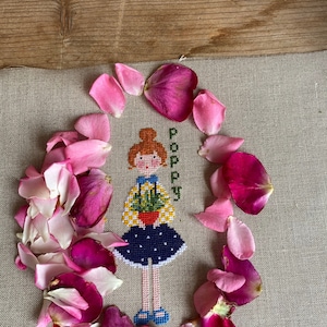 May include: Cross-stitch embroidery of a girl holding a potted plant, surrounded by pink rose petals on a beige linen fabric. The girl is wearing a yellow and white striped shirt, a blue skirt with white polka dots, and blue shoes. The embroidery is titled "Poppy".