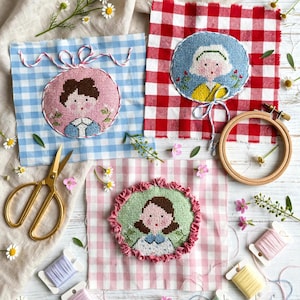 Può includere: Tre ritratti a punto croce su quadrati di tessuto a quadretti vichy blu, rossi e rosa. I ritratti presentano dettagli ricamati, tra cui un cerchio rosa con spago rosso e bianco, un cerchio rosso con spago blu e un bordo rosa con volant.