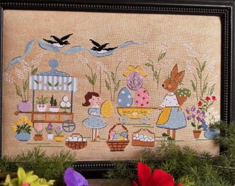 Könnte beinhalten: Gerahmte Kreuzstich-Stickerei, die eine Ostermarktszene darstellt. Ein Mädchen und ein Hase verkaufen Eier und Blumen. Das Kunstwerk zeigt ein blaues Band mit zwei schwarzen Vögeln, Eierkörbe und bunte Blumen.